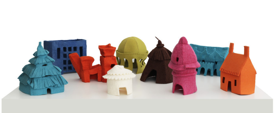 A set of nine miniature houses made from felt sit on a white gallery display stand in front of a white background. These small sculptures each a different color and serve as simplified representations architectural traditions from around the world.