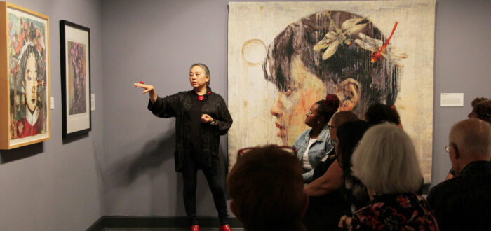 Hung Liu speaks to an audience about her paintings of Chinese women, which hang on the walls around her. Liu is a light-skinned adult woman with gray hair in a low ponytail, dressed in black with red jewelry and shoes. She stands, gesturing with one hand at a painting to her right.