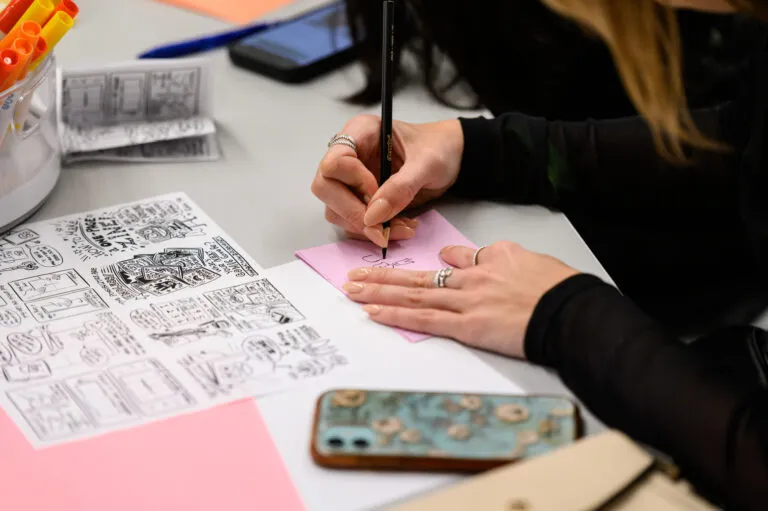 A person draws on a pink sheet of paper with another piece of paper with instructions alongside.